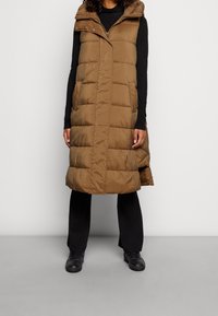 Kobieta ubrana w bezrękawnik w brązowym kolorze na czarną bluzkę z długim rękawem, czarne spodnie i czarne buty, stojąca na tle jednolitego tła.