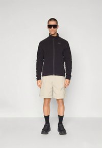 Άνδρας που φοράει μαύρο φερμουάρ, μπεζ σορτς, μαύρες κάλτσες, μαύρα αθλητικά παπούτσια και μεγάλα σκούρα γυαλιά ηλίου, στέκεται μπροστά από απλό φόντο.