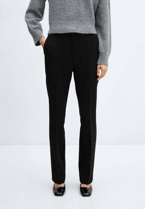 Kobieta ubrana w czarne, dopasowane spodnie, szary krótki sweter i czarne lakierowane buty z kwadratowym noskiem, stojąca z jedną ręką w kieszeni.