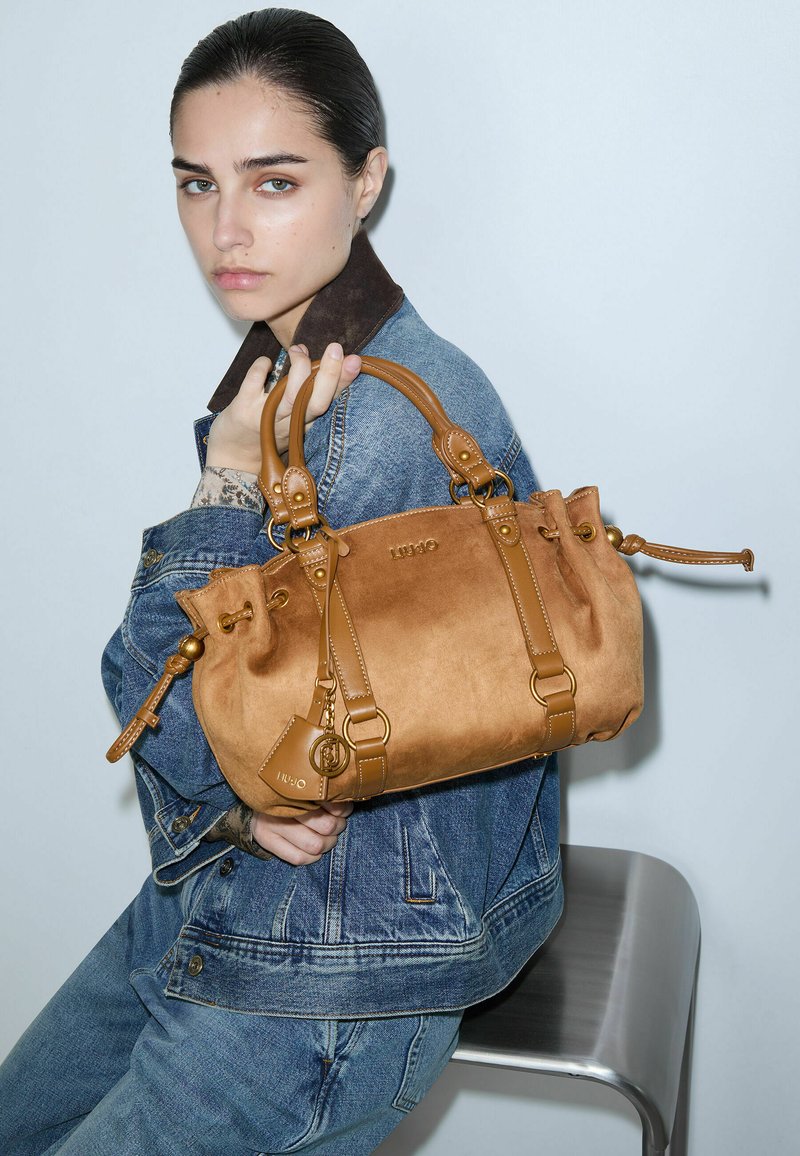 Jeune femme en veste en jean et jean tient un sac à main en cuir marron tout en étant assise sur un tabouret en métal contre un mur uni.