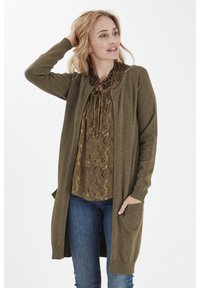 Olive roheline kudumiskardigan pika disainiga, soonikute äärtega ja avatud esiosaga. Kandestatud mustrilise pruun blousee peale, koos siniste teksadega.