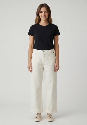 Femme debout, portant une chemise noire à manches courtes, un pantalon large crème taille haute avec ourlets retournés, et des baskets beige, sur un fond uni.