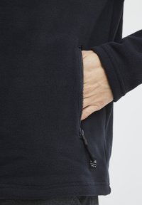 Temno modra flis jakna z stranskim žepom na zadrgo, gladko teksturo in majhno oznako na robu. Ohlapna zasnova, dolgi rokavi.