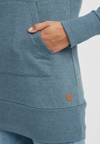 Sudadera de manga larga en color azul verdoso con un bolsillo tipo canguro, puños de canalé y un pequeño parche de logo marrón cerca del dobladillo. Textura suave.