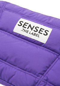 Lila Puffjacke mit horizontalem Steppmuster. Auf der Vorderseite befindet sich ein weißes rechteckiges Etikett mit der Aufschrift "SENSES .THE LABEL", das aufgenäht ist.