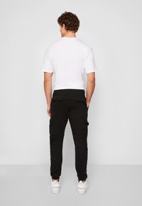 Pánský outfit zahrnující bílou polokošili s krátkým rukávem a černé cargo kalhoty s bočními kapsami, doplněné bílými teniskami.