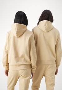 Sudaderas beige de tejido suave, con bolsillo tipo canguro y puños acanalados, llevadas con pantalones de chándal a juego. Vista desde la parte de atrás.