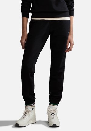 Personne portant un pantalon de jogging noir, des chaussettes blanches avec des accents verts et des baskets montantes blanches, debout devant un fond uni.