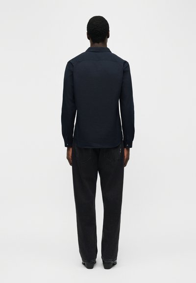 Mann trägt ein dunkelblaues, geknöpftes Hemd und lockere schwarze Jeans, steht mit dem Rücken zur Kamera vor einem schlichten weißen Hintergrund.