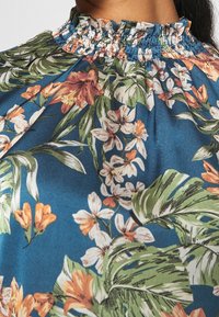 Blouse fleurie avec un col froncé, présentant un fond bleu et des fleurs orange vif et blanches avec des feuilles vertes. Texture de tissu lisse.