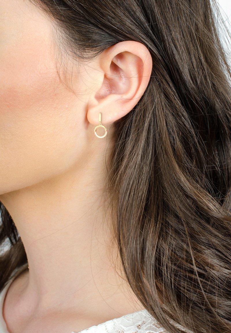 Orecchini a cerchio in oro con superficie testurizzata, dotati di un design a goccia e un post dritto, indossati all'orecchio di un modello con capelli di lunghezza media.