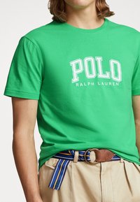 Zelené bavlněné tričko s bílým potiskem "POLO RALPH LAUREN". V kombinaci s khaki kalhotami a pruhovaným opaskem s modrými, žlutými a hnědými akcenty.