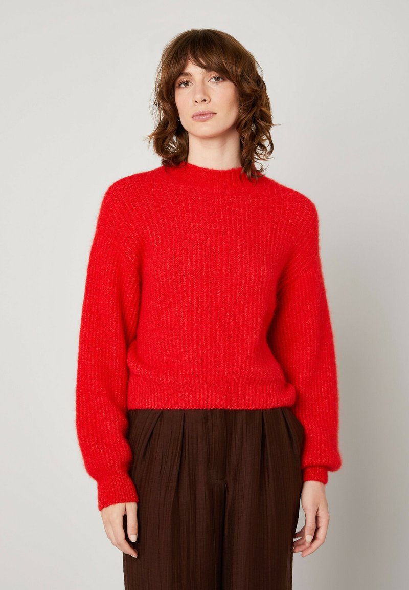 Raudonas megztinis su aukštu kaklu, su raukšlėtu paviršiumi ir ilgomis rankovėmis. Dėvimas ant rudos klostuotos kelnės.