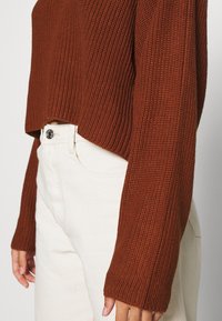 Maglione corto color ruggine con texture a coste e maniche lunghe, abbinato a jeans chiari a vita alta con dettaglio dei bottoni.