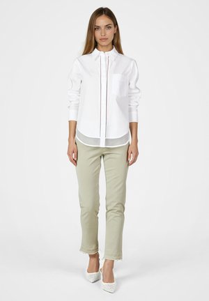 Junge Frau steht, trägt weißes Hemd mit Knöpfen, beige Hosen mit ausgefransten Säumen und weiße spitze Absätze, vor einfachem Hintergrund.