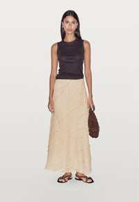 Top sans manches noir avec une jupe maxi beige texturée présentant des lignes ondulées. La tenue est complétée par des sandales noires et un sac en perles marron.