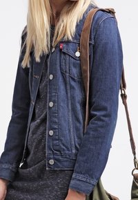 Mörkblå denimjacka med metallknappar, bröstfickor med logotyp och skräddarsydd passform. Bärs över en mörk melerad klänning.