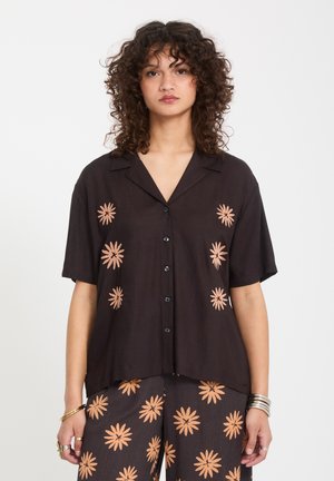 Femme aux cheveux bouclés portant une chemise noire boutonnée et un pantalon assorti avec des motifs de fleurs pêche, debout devant un fond blanc.