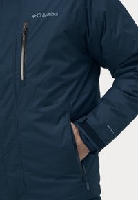 Marineblaue Columbia-Jacke mit Reißverschlusstasche auf der Brust, verstellbaren Bündchen und Omni-Tech-Branding. Glatte Textur, isoliertes Design.