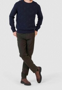 Homme portant un pull texturé bleu marine, un pantalon vert foncé et des bottes en cuir marron, debout avec les jambes croisées devant un fond uni.