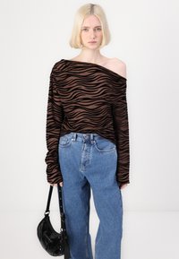 Smeđi i crni prugasti dugi rukav top s off-shoulder dizajnom, uparen s visokim plavim trapericama i crnom torbom.