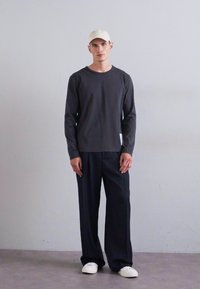 Man i beige keps, mörk långärmad skjorta, lösa svarta byxor och vita sneakers står mot en enhetlig ljusgrå vägg.