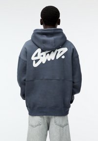 Modrá mikina s kapucí s velkým bílým grafickým nápisem na zadní straně. Design zahrnuje uvolněný střih a žebrované manžety.