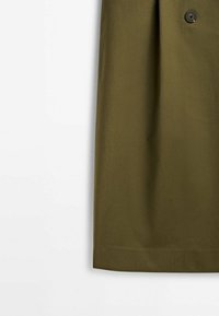 Gros plan sur la partie inférieure d'une jupe vert olive avec un détail de bouton sur le côté droit sur fond blanc.