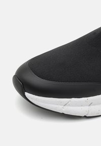 Czarne tenisówki z teksturowaną górą z tkaniny, gumowym czubkiem i białą, nakrapianą podeszwą środkową. Wygięty kształt z gładkimi akcentami.