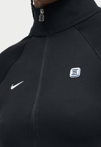 Κοντινό πλάνο ενός ατόμου που φοράει μαύρο αθλητικό τζάκετ με φερμουάρ, με το λογότυπο της Nike στο αριστερό στήθος και ένα μικρό τετράγωνο έμβλημα στο δεξί στήθος.