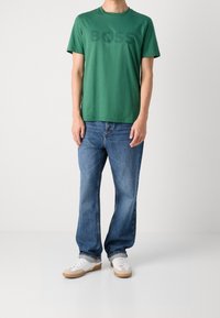 Henkilö, joka pitää yllään vihreää T-paitaa, jossa on "BOSS"-teksti, sinisiä farkkuja ja valkoisia lenkkareita, joissa on kumipohjat, seisoo vaaleaa taustaa vasten.