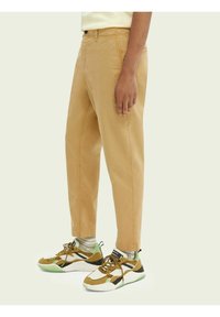 Khaki Chinos im geraden Schnitt, mit Seitentaschen, Gürtelschlaufen und einem umgeschlagenen Saum, kombiniert mit mehrfarbigen Sportschuhen.
