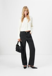 Woman wearing white buttoned cardigan, black wide-leg jeans, black pointed heels, and holding a black handbag with braided handles.