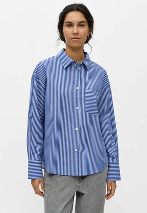 Femme portant une chemise à rayures verticales bleu et blanc avec une poche et un jean gris, debout devant un fond blanc uni.