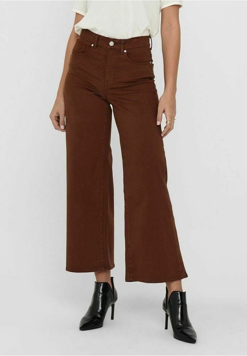 Vrouw in hoge taille, brede bruine broek, met een wit ingestopt topje en zwarte puntige hoge hakken, staande op een grijze vloer.