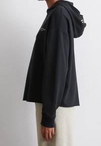 Zwarte hoodie met lange mouwen, relaxte pasvorm en logo detail aan de voorkant. Zachte stof met een gladde textuur, gecombineerd met lichtgrijze onderkleding.