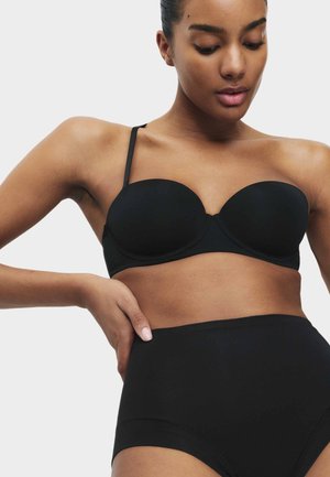 Frau trägt einen schwarzen trägerlosen BH mit abnehmbaren Trägern und hoch taillierte schwarze Slips, schaut nach unten und hat eine Hand auf der Hüfte.