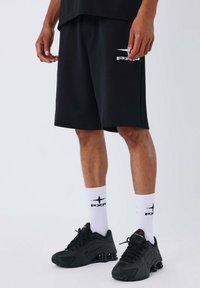Shorts de sport noirs en tissu doux, avec un logo blanc. Accompagnés de chaussettes blanches affichant un logo noir et de baskets noires.