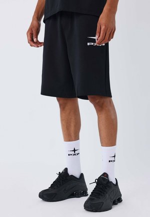Pantalones cortos deportivos negros de tela suave, con un logo blanco. Combinados con calcetines blancos que tienen un logo negro y zapatillas negras.