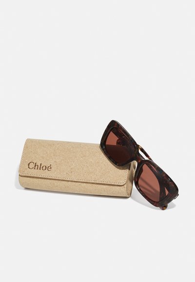 Occhiali da sole tartarugati con lenti scure accanto a un astuccio in sughero beige con logo "Chloé" in rilievo e design a pieghe.