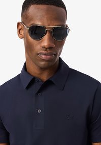 Hombre con gafas de sol oscuras y una camiseta tipo polo azul marino con un pequeño logo de cocodrilo bordado sobre un fondo blanco.