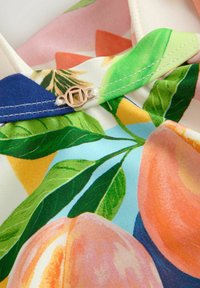 Tissu multicolore présentant des motifs de pêches et de feuilles, avec un col bleu contrastant et un accent logo doré au niveau de l'encolure.