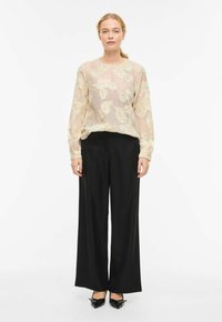 Blouse beige clair transparente avec broderie florale, manches longues, associée à un pantalon ample noir et des chaussures noires à bout pointu.