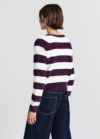 Person mit kurzem braunem Haar, die silberne Creolen trägt, einen lila und weißen gestreiften Pullover sowie weite dunkelblaue Jeans trägt und seitlich steht.