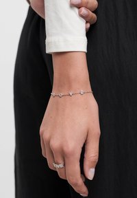 Sølvarmbånd med "KÆRLIGHED" bogstaver prydet med små klare krystaller, båret på en håndled med en hvid langærmet top, mod en mørk baggrund.