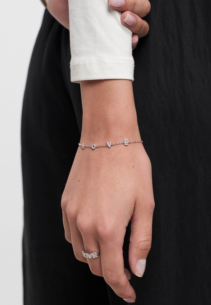 Sølvarmbånd med "KÆRLIGHED" bogstaver prydet med små klare krystaller, båret på en håndled med en hvid langærmet top, mod en mørk baggrund.