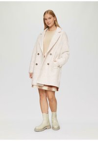 Doppelreihiger cremefarbener Mantel mit übergroßen Revers, strukturiertem Finish und seitlichen Taschen, kombiniert mit einem gemusterten Rock und beigen Ankle-Boots.