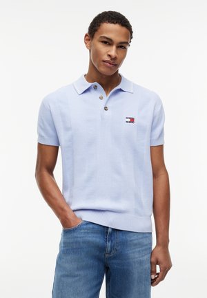 Młody mężczyzna ubrany w jasnoniebieską dzianinową koszulkę polo z krótkim rękawem, guzikami i logo, w połączeniu z niebieskimi dżinsami, na białym tle.