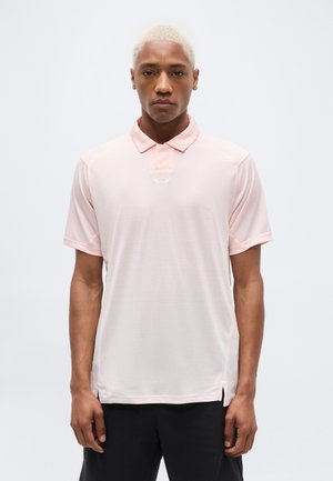 Uomo con capelli biondi corti che indossa una polo rosa chiaro e pantaloni neri, in piedi davanti a uno sfondo bianco semplice.