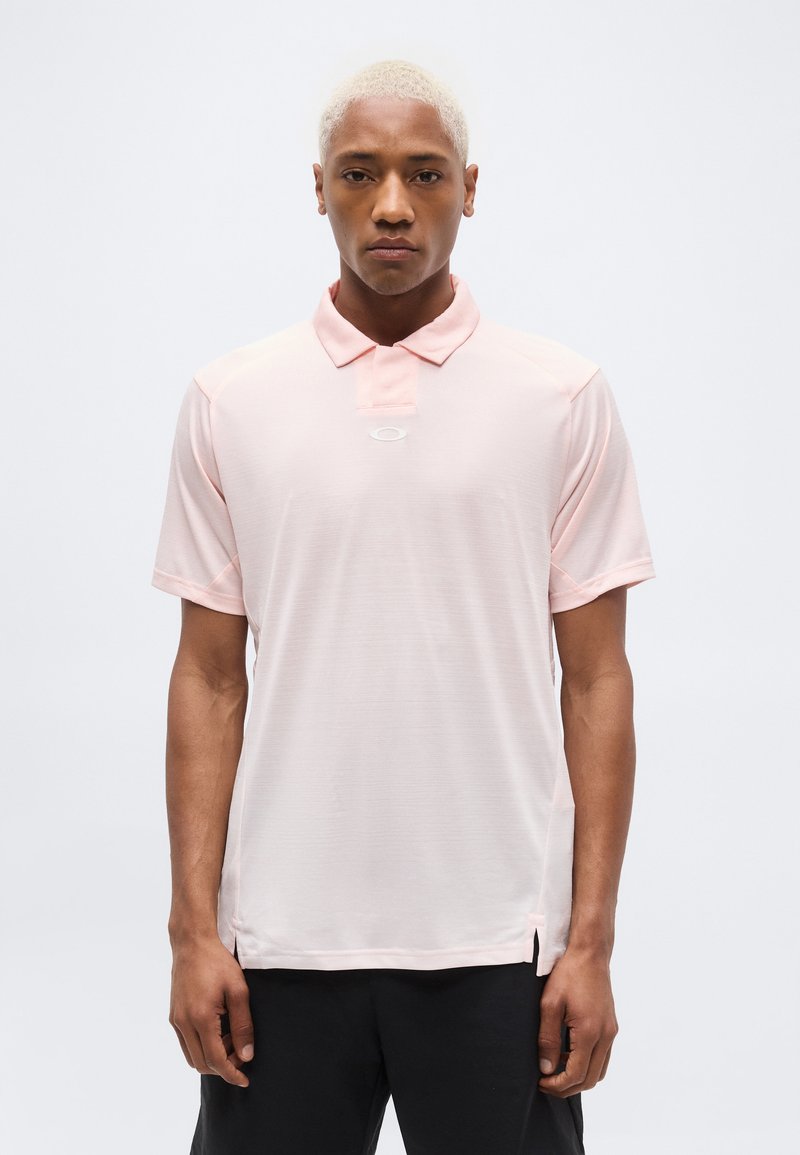 Άνδρας με κοντά ξανθά μαλλιά, φοράει ανοιχτό ροζ πόλο μπλουζάκι και μαύρο παντελόνι, στέκεται μπροστά σε απλό λευκό φόντο.